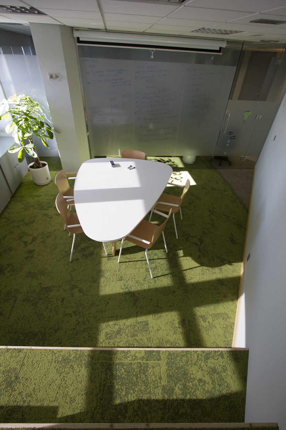 Vista superior de la sala de reuniones de una oficina en Tetuán, Madrid, con moqueta verde, mesa blanca y pared de vidrio para escribir