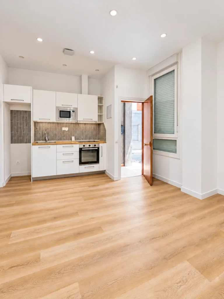 Estudio terminado con cocina blanca al fondo, suelo de madera clara y una ventana alta
