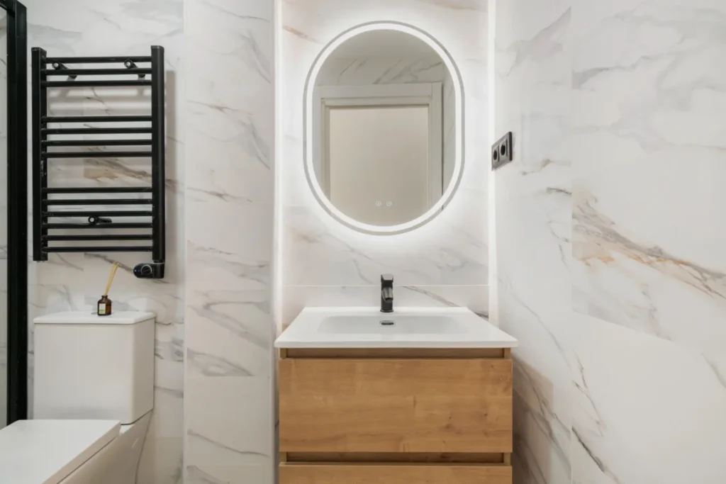 Baño con paredes de mármol blanco, mueble de madera y espejo ovalado iluminado