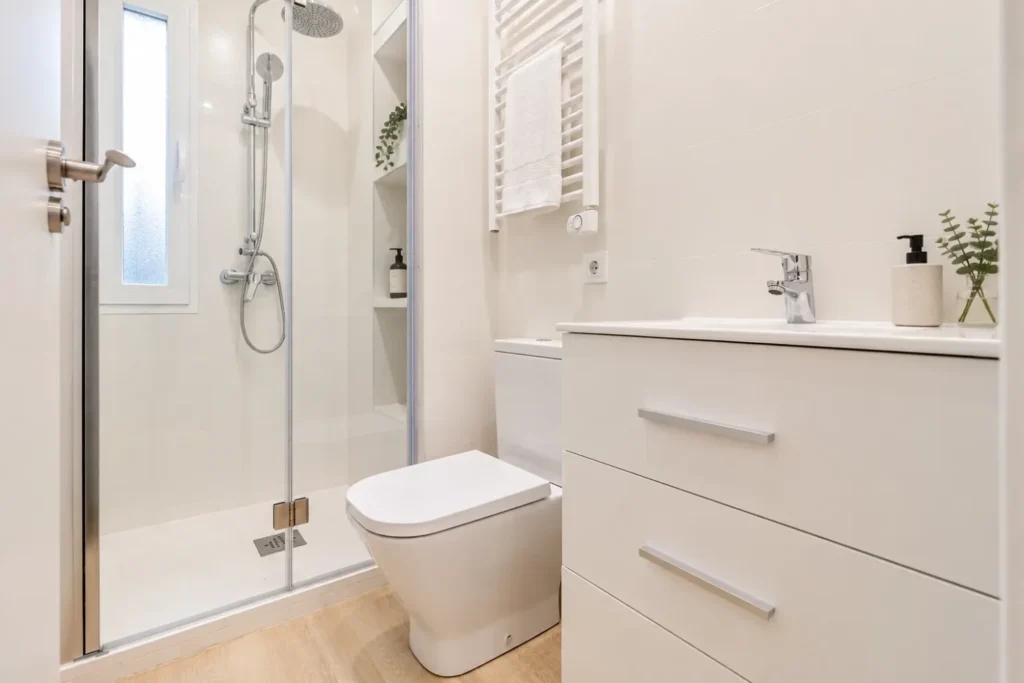 Baño blanco con ducha, mampara de vidrio y mueble de lavabo con cajones