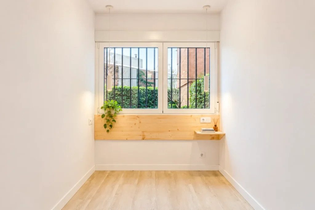 Estancia blanca con suelo de madera clara, ventana con reja y una repisa con una planta