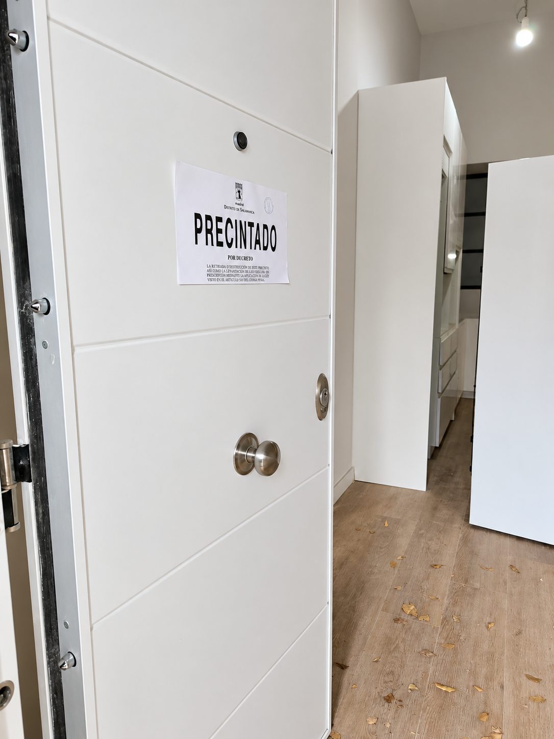 Puerta blanca de una vivienda entreabierta con un cartel de «Precintado» pegado, y al fondo un interior en obras con muebles de cocina sin terminar