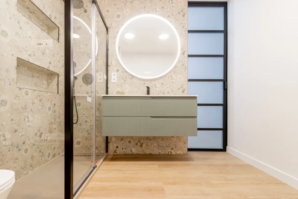 Baño con mueble de lavabo verde, espejo redondo iluminado y mampara de vidrio