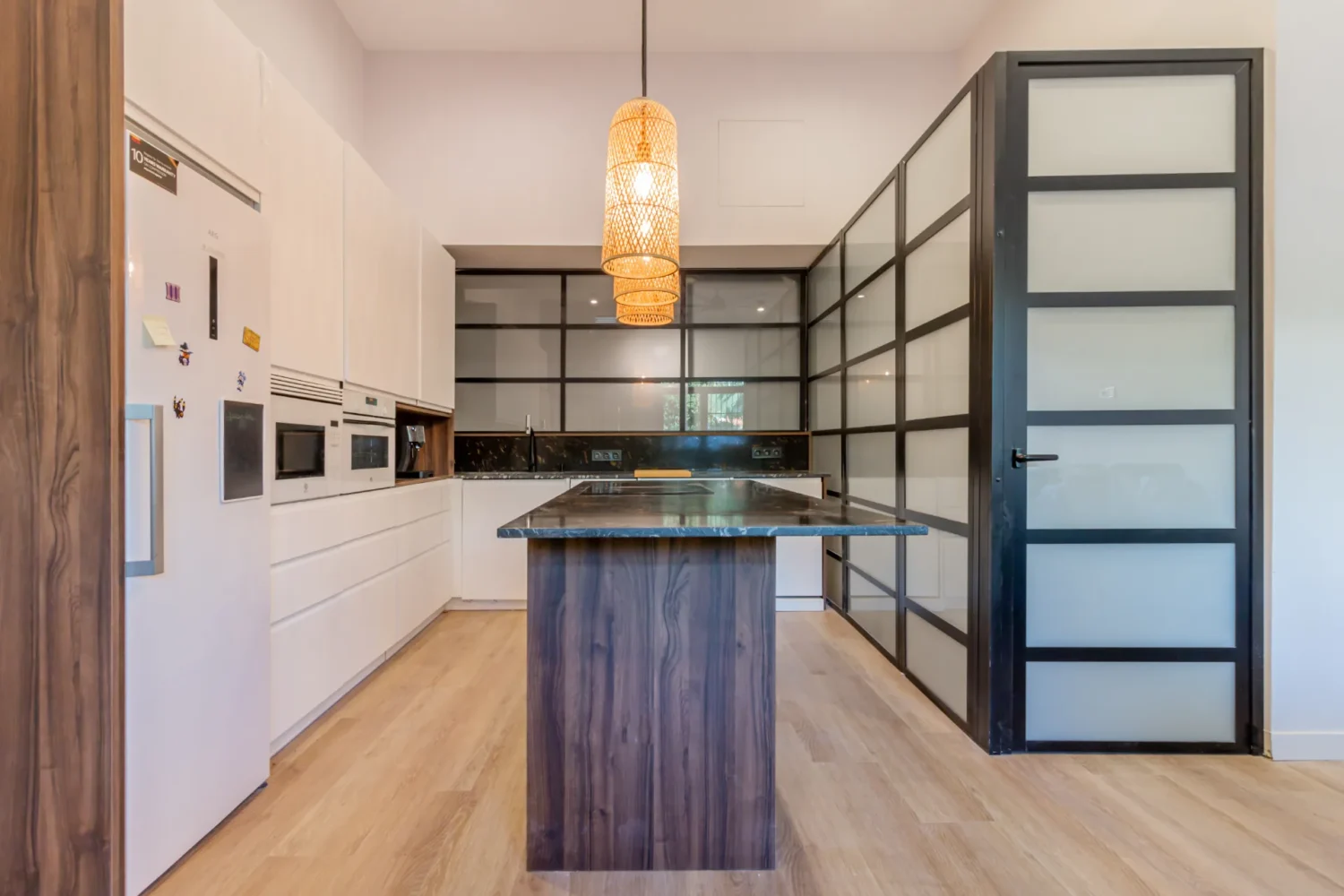 Cocina blanca con isla de madera oscura, lámpara colgante de mimbre y cerramiento acristalado con perfil negro