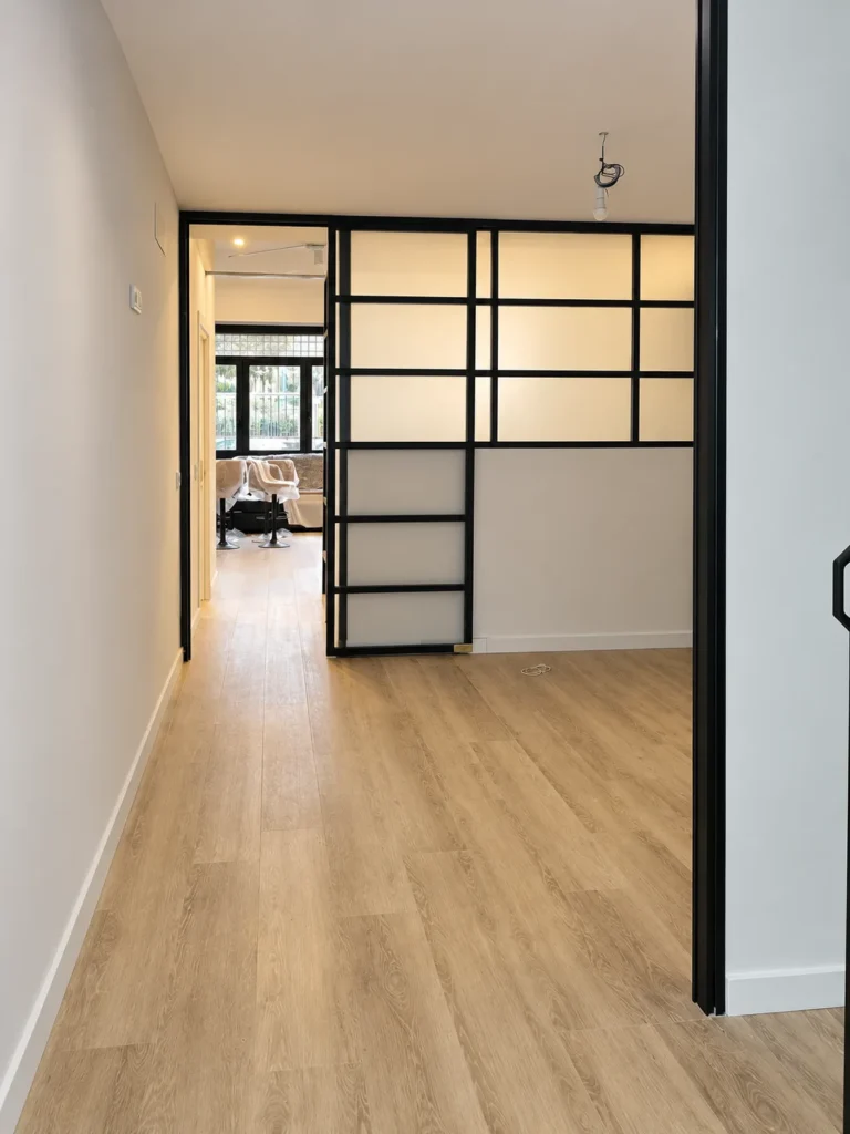 Pasillo con suelo de madera clara que termina en una puerta corredera de vidrio con perfil negro