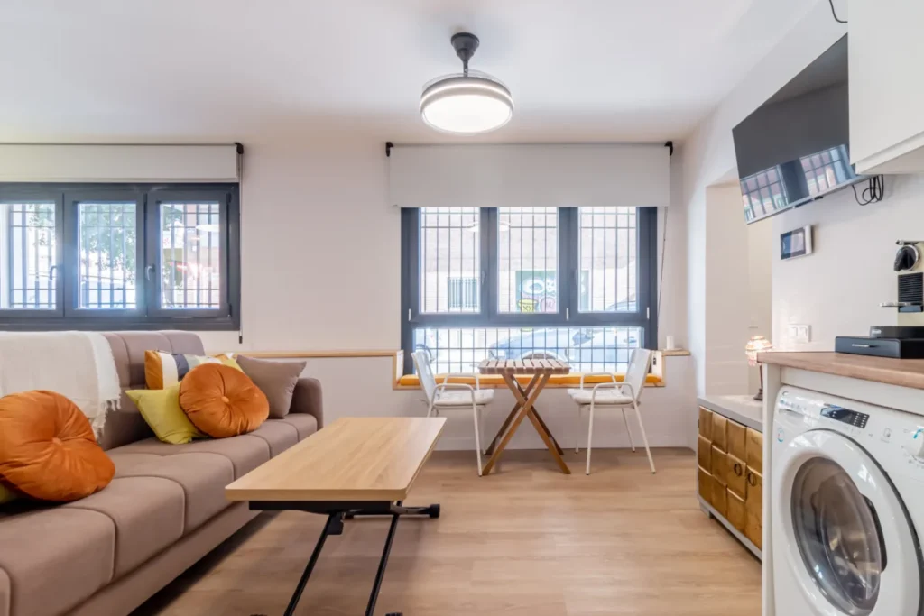 Salón con sofá gris y cojines naranjas, mesa de madera y ventanas con rejas al fondo
